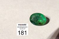 Close-up photo of a translucent oval faceted emerald gemstone showing natural inclusions and green color, placed on white background next to auction lot number.