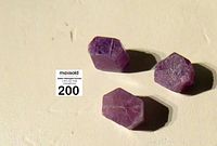 Three natural six-sided crystals in purple hues placed on a light surface with auction tag visible.