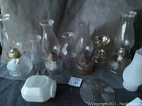 Four complete kerosene lanterns displayed in a row with glass chimneys, metal and glass bases, plus extra kiln chimneys and parts laid in front; visible signs of light use and age.