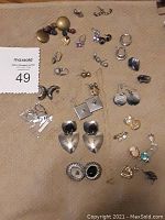 Overhead image showing a variety of earrings and necklace charms laid out on a beige surface. Includes assorted gold and silver tones in different styles and sizes.