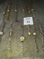 Overview of 12 ladies' silver-tone linked band wristwatches arranged on a flat surface showing diversity in dial colors, face shapes, and some turquoise stones on one band.