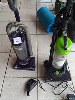 Photo showing the purple and silver Eureka upright vacuum and the black and green Bissell upright vacuum on tile floor with yoga mat in back.