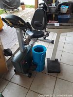 Photo showing the Schwinn exercise bike with black seat and pedals next to a rolled blue exercise mat and the vintage medical scale on tiled floor.