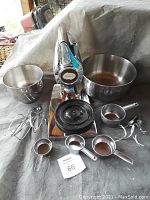 Photo showing Sunbeam Mixmaster mixer with two mixing bowls, four metal measuring cups, and two types of beaters arranged around the mixer on a grey cloth surface.