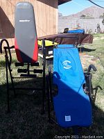 Photo showing the BodyFit inversion bench with red and black padded backrest, metal frame and foam leg rollers next to the blue AB Chair glider in a grassy outdoor area.