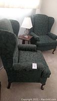 Pair of dark green upholstered wing back chairs shown with fabric floral pattern, wooden legs, and padded armrests.