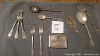 Photo of a collection of silver utensils and a small decorative silver box on a grey cloth background. Includes forks, spoons, servers.