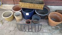 Multiple assorted planting pots of various materials including ceramic, plastic, bamboo, and metal, arranged side by side on a paved surface with a brick wall background.