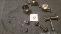 Photo of silver sugar and creamer set, ribbed cup, metal tongs, baby rattle, and small pierced shaker item placed on a cloth.