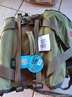 Top view showing olive green pannier bag with two brown adjustable straps and buckles, still has attached tag with product information and auction label.