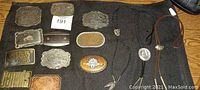 Top view showing 12 belt buckles laid out on a black cloth next to three bolo ties with different types of cords and two pendants.