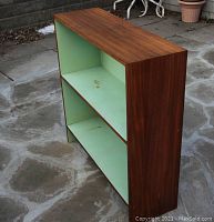 Stand-alone teak bookshelf shown at an angle revealing the side grain and light green interior shelves with visible stains and wear.