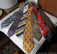 7 narrow silk ties laid out on a white cloth, showing various colorful patterns and designs.