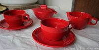 Red Melmac cups, saucers, sugar bowl with lid, and creamer arranged on a cloth surface showing front angles and details of handles and lids.
