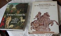 Two large hardcover art books placed side by side on a white cloth surface: 'Lucas Cranach' on the left with color dust jacket, 'Masterpieces of Western Sculpture' on the right with white dust jacket and sculpture photo