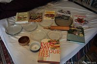 Overview of lot including Pyrex pie plates arranged in a group, cookbooks placed behind them, rolling pin lying horizontally, and ceramic forms at front.