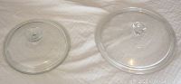 Two Pyrex glass pot lids side by side on a light-colored cloth background, showing clear glass with knob handles.