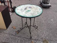 Full view of round outdoor patio table with mosaic bird and floral ceramic top and iron scrollwork legs on asphalt surface.
