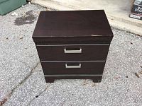 Full view of modern dark brown wooden two drawer night stand with metal handles showing some wear on top surface