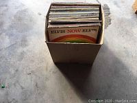 Photo of a cardboard box containing about 50 vintage LP record albums including "Elvis Now" by Elvis Presley visible at front.