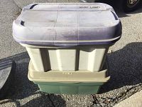 Two stacked plastic storage bins outdoors on asphalt. Top bin gray lid marked 'XMAS LIGHTS', bottom bin green with beige lid.