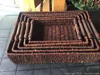 Five stacked rectangular wicker basket trays with wooden handles in dark brown, viewed from side angle showing nested sizes.