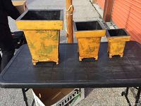 Set of three yellow metal planters with green rooster illustrations placed on a black table outdoors.