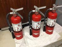 Photo showing three red Amerex fire extinguishers standing upright on a white towel against a dark wood background.