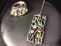 Close-up image of the ring and necklace pendant on a black leather surface showing the textured silver and various colored stones.