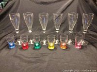 Front view of 6 clear pear-shaped wine glasses and 6 colorful shot glasses arranged in front on black fabric background.