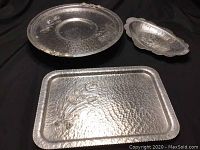 Full set of three hand wrought metal serving pieces by Rodney Kent on black backdrop, showing round pedestal platter, oval platter and rectangular tray.