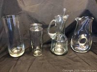 Four clear glass items on black cloth background: two tall vases on the left and two glass pitchers on the right, one pitcher with glass stir stick inside.