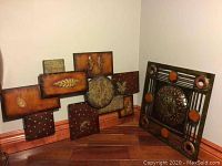 Photo showing two pieces of metal wall art placed against a light wall on a wooden floor. One piece is large with multiple embossed panels featuring nature motifs in brown tones. The other piece is a square with a central circular textured pattern surrounded by orange circles and linear rods.