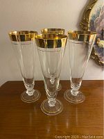 Front-facing photo showing all four tall liquor glasses with clear glass and thick gold rims, set on wooden surface