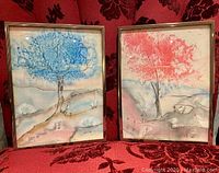 Photo showing both framed paintings side by side on fabric background, highlighting blue and red tree artwork and frame wear.