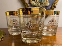 Four vintage drinking glasses with thick gold rims and gold atomic-era motifs arranged on wooden surface, showing front and side views.
