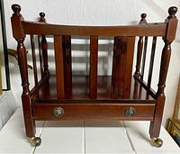 Front view of vintage wooden magazine rack showing open storage area, drawer with metal pull, and brass wheels.