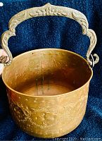 Front angle of antique hammered copper bucket showing embossed floral patterns and ornate brass handle with floral engraving.
