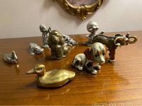 Nine miniature metal animal figurines on wooden surface including brass duck and painted elephant