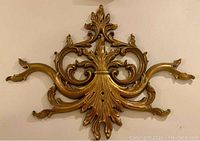 Front view of gold tone resin wall decor showing symmetrical ornate scroll and leaf motif, some wear visible.