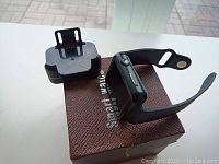 Side angle of black smart watch on brown textured box with charging dock showing the strap and charging pins.