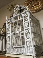 Large white wicker birdcage shown standing upright, featuring rounded dome top with hanging ring, front door with wicker latch and ornate scrolling woven pattern.