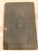 Front cover of the antique book shows textured and embossed dark green cover with intricate central design and some wear.