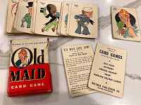 Top-down view of playing cards fanned out showing cartoon characters, instruction cards, Old Maid card, and original box.