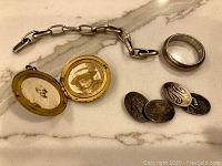 Photo showing gold-tone locket open displaying two black and white photos inside, silver-tone chain, silver-tone band ring, and four oval cufflinks with engravings.