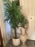 Photo showing full artificial plant with multiple green leaves and white planter beside ornate urn against white door background.