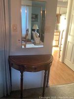 Half round wooden side table with drawer below wall mirror reflecting room interior.