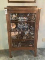 Front view of vintage wooden curio cabinet with glass door displaying silver-plated serving items on three shelves.