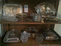Shelf with various silver-plated trays, covered serving dishes, round trays, and clear glass/crystal bowls and sugar bowl.