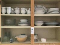 Kitchen cabinet with shelves containing white mugs with grey patterns, white plates and bowls, clear glass bowls, and a large tan glass casserole dish with lid